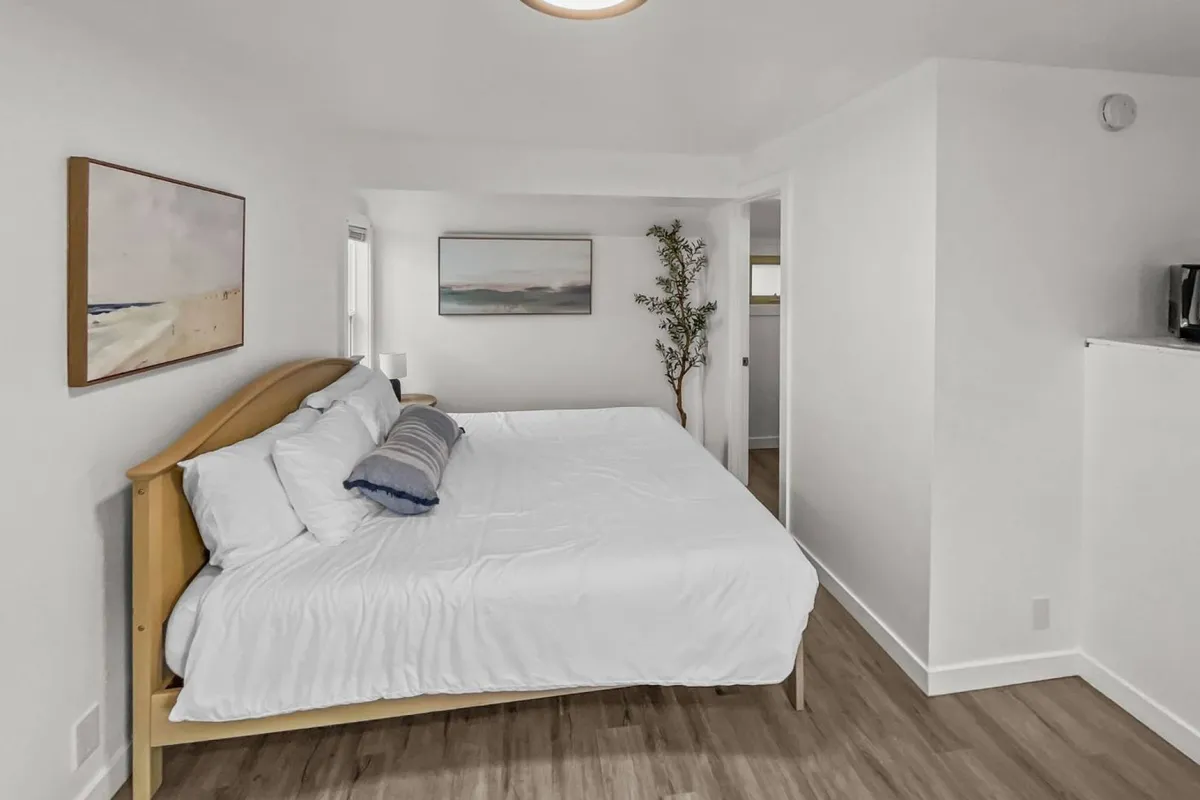 A cozy interior guest room featuring a comfortable bed adorned with neutral linens, a potted plant adding a touch of greenery, and artistic wall decor. Natural light pours in through a nearby window, offering a tranquil and welcoming space for relaxation and quiet conversation.