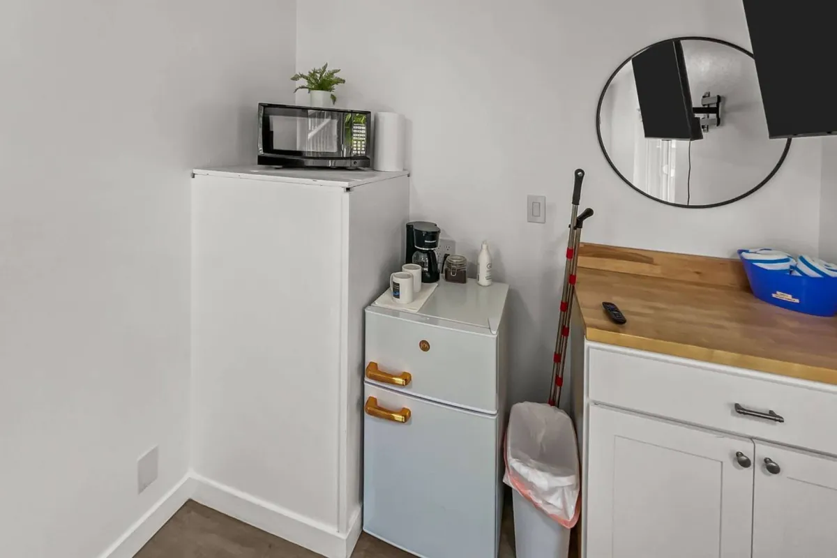 A kitchenette area showcasing a compact refrigerator, microwave, and kitchen countertop, all styled in a modern, inviting manner. A round mirror adds visual interest and reflects the thoughtful design, making the space feel more open while highlighting the studio's convenience for preparing snacks or simple meals.