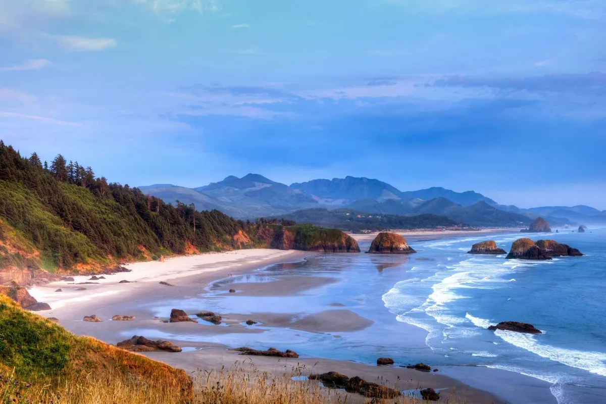 A stunning view unfolds across the sandy beach, where waves meet the shore and dramatic rock formations—including Twin Rocks Arch—rise against the sky. Distant mountains and green forest complete the backdrop, evoking the magic and natural beauty of the Oregon Coast.