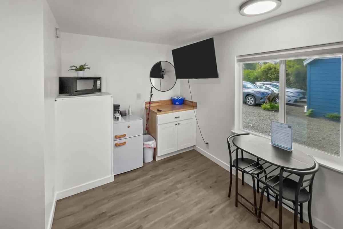 The hero image showcases a bright, inviting interior with a well-equipped kitchenette featuring a microwave and refrigerator, a warm wooden counter perfect for casual meals, and a cozy seating area with a television. A window offers a glimpse of the coastal scenery, suggesting the unit’s proximity to the beach.