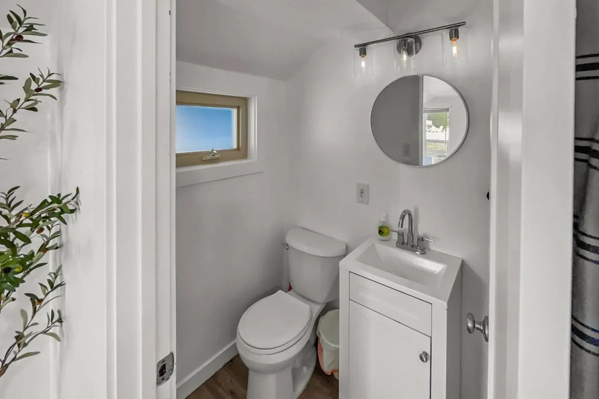 A refreshing and minimalistic bathroom scene featuring a toilet, sink with mirror, window allowing natural light, and small decorative plants. The setup conveys a sense of cleanliness and comfort, ideal for personal grooming and relaxation.