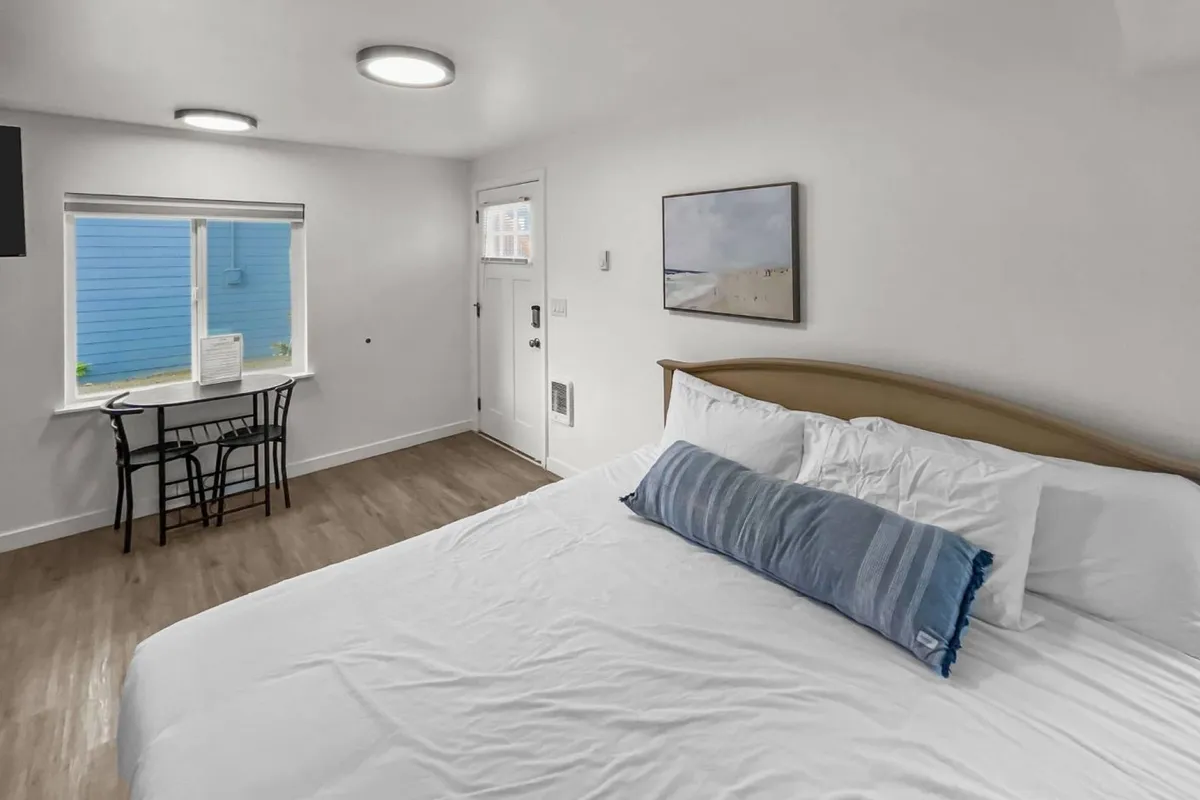 A sunlit studio interior featuring a comfortable king-size bed with a blue accent pillow, a window that lets in natural light and offers a view outside, and a small table with chairs perfect for relaxing, reading, or enjoying coffee. The modern furnishings and open-concept layout are evident throughout.